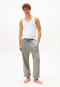 Junger Mann barfuß in einem weißen Tanktop und grauen Sweatpants, der vor einem schlichten weißen Hintergrund steht.