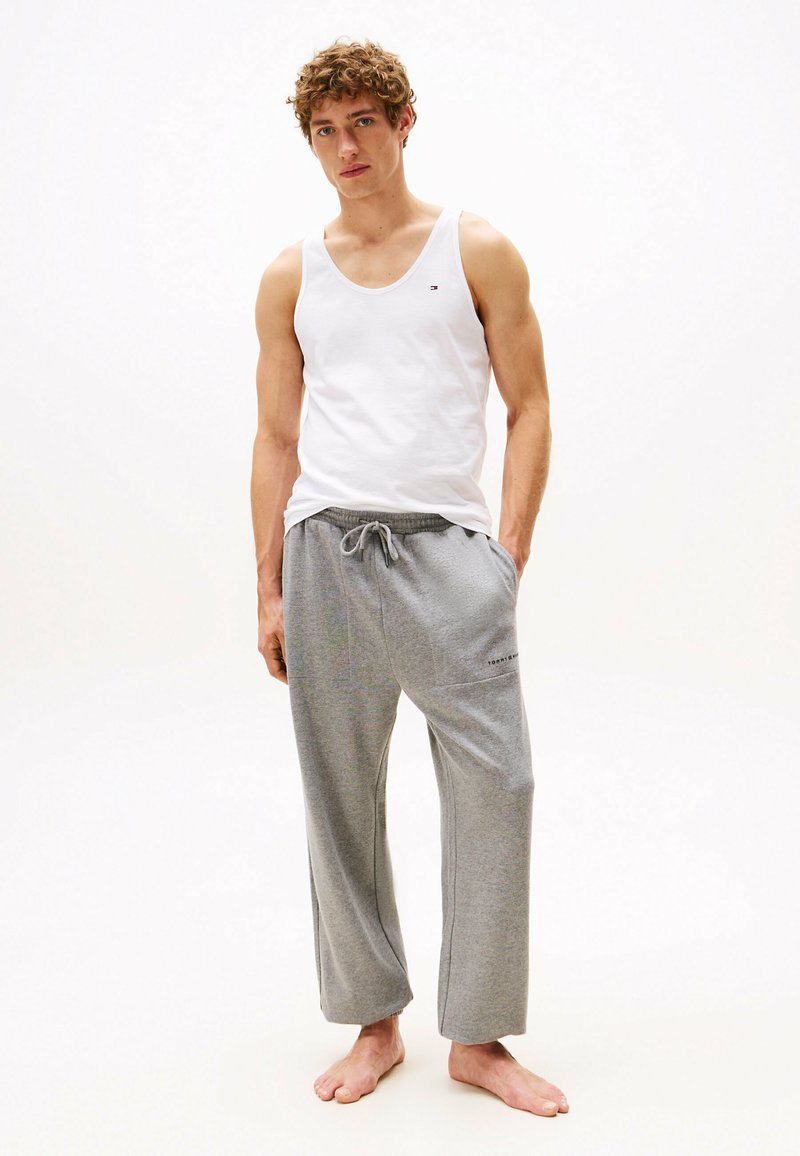 Junger Mann barfuß in einem weißen Tanktop und grauen Sweatpants, der vor einem schlichten weißen Hintergrund steht.