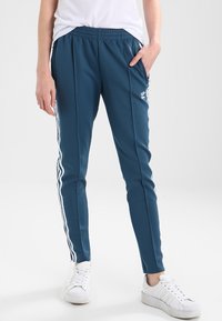 Tealfärgade träningsbyxor med vita sidostrek, elastisk midja och fickor. Matchas med vita sneakers på en neutral bakgrund.