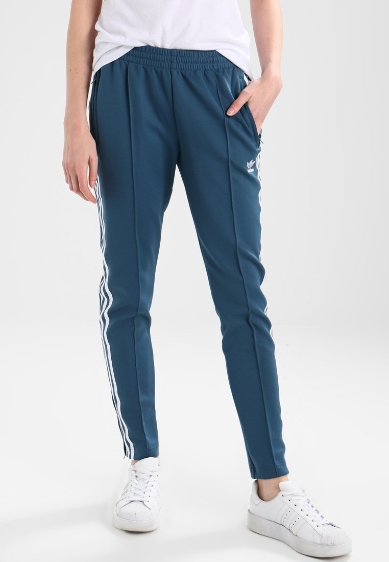 Tealfärgade träningsbyxor med vita sidostrek, elastisk midja och fickor. Matchas med vita sneakers på en neutral bakgrund.