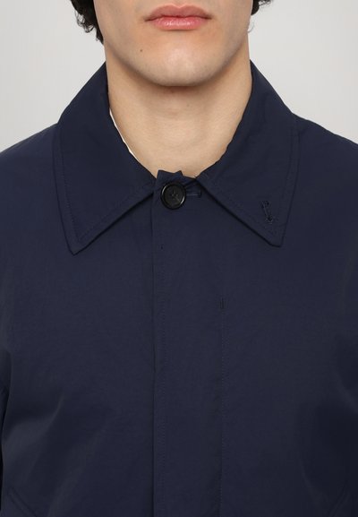 Veste bleu marine avec un grand col, fermeture à un bouton et tissu texturé. Présente des coutures surpiquées et un minimalisme dans les détails.