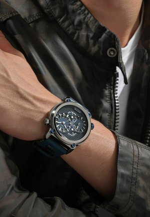 Blau- und silberne Uhr mit einem runden Metallgehäuse, strukturiertem Zifferblatt, doppeltem Zeitanzeigefeld und einem Gummiband, getragen am Handgelenk.