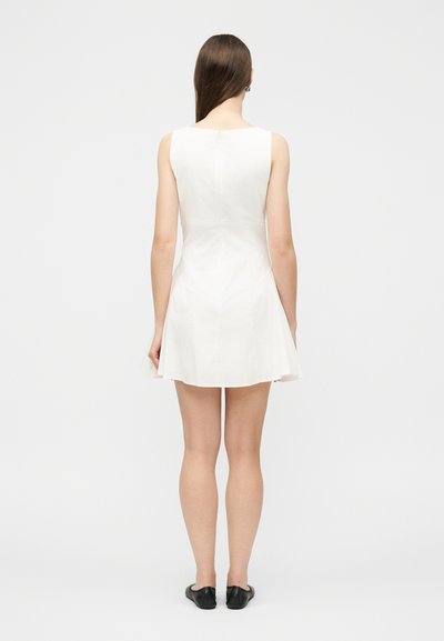 Γυναίκα με μακριά καστανά μαλλιά, ντυμένη με λευκό μίνι φόρεμα χωρίς μανίκια και μαύρα παπούτσια με επίπεδη σόλα, στέκεται με την πλάτη προς τα εμπρός σε απλό φόντο.