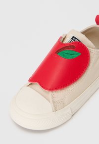 Accent de chaussure en toile beige en forme de pomme en cuir rouge avec un embout en caoutchouc et des détails cousus. Design minimaliste avec des éléments ludiques.