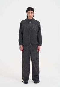 Schwarze Zip-Jacke mit geprägtem Text, kombiniert mit passenden lockeren Hosen mit Cargo-Taschen. Der Stoff wirkt strukturiert und langlebig.