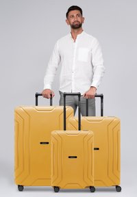 Tre valigie rigide gialle di dimensioni diverse con superfici texturizzate, maniglie nere retrattili e ruote per la mobilità.