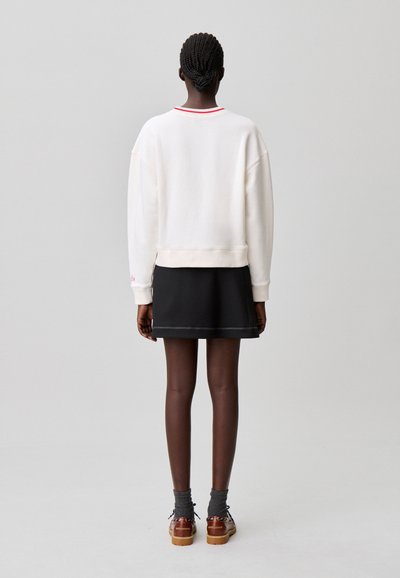 Sweat-shirt blanc avec bordure rouge, coupe décontractée et poignets côtelés, associé à une jupe noire. Présentant un design minimaliste et une texture lisse.