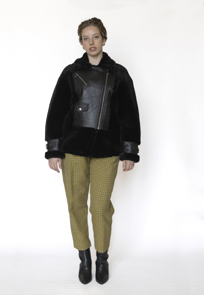 Zwarte leren jas met nepbont en een ritssluiting zak, gecombineerd met gele geruite broek en zwarte enkellaarsjes met hak. Effen achtergrond.