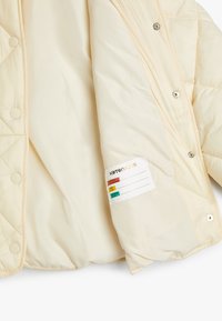 Ljust beige quiltad pufferjacka med tryckknappar och en etikett på ärmen som anger storlek och skötselråd. Mjuk, slät textur.