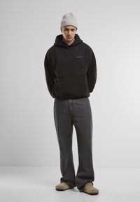 Schwarzer Hoodie mit Fronttasche und dezentem Logo, kombiniert mit dunkelgrauen, weit geschnittenen Jeans und beige-farbenen Slippern. Strukturierte, gerippte Strickmütze.