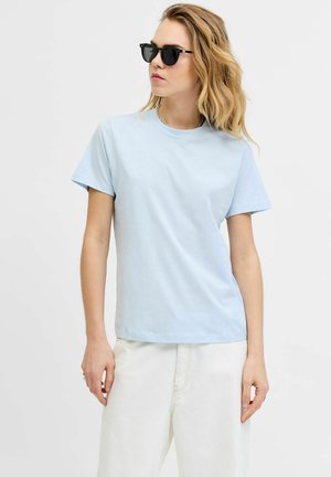 Femme portant des lunettes de soleil noires, un t-shirt bleu clair à manches courtes et un pantalon blanc, debout devant un fond clair uni.