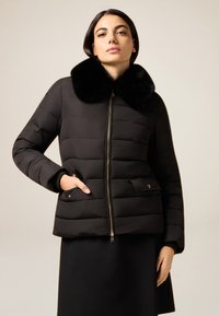 Schwarze Steppjacke mit Kunstpelzkragen, ausgestattet mit Reißverschluss vorne, gesteppter Optik und zwei Seitentaschen. Glatte Textur, mittlere Länge.