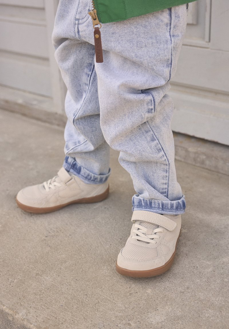Kind trägt hellblaue Jeans mit Umschlägen und beige Sneaker mit Klettverschluss, steht auf Beton.