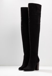 Botas negras por encima de la rodilla hechas de suave ante, con punta afilada y taco cuadrado con textura de madera. Detalle de cremallera en el lateral.