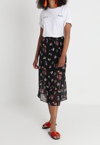 Nainen, jolla on päällään valkoinen "ÄLÄ ODOTA" -t-paita, musta kukkakirjailtu midihame, oranssit tupsukorvakorut ja punaiset liukupohjakengät, seisoo valkoista taustaa vasten.