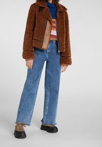 Ruskea tekoturkistakki vetoketjulla edessä, yhdistetty sinisiin leveälahkeisiin farkkuihin ja ruskeisiin nauhallisiin kenkiin, joissa on mustat pohjat.