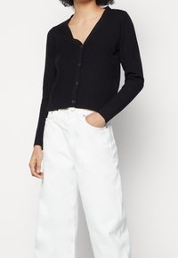 Cardigan negro acanalado con escote en V, mangas largas y cierre de botones en el frente. Combinado con pantalones vaqueros anchos de tiro alto en blanco.