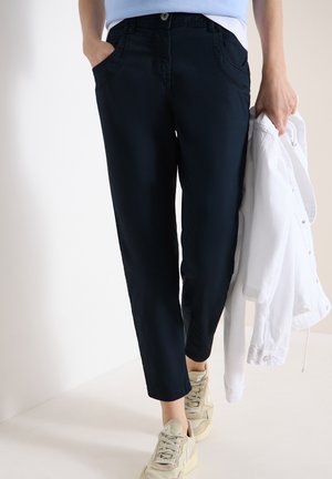 Kvinde iført sorte, tilspidsede bukser og beige sneakers, der holder en hvid jakke i den ene hånd og har den anden hånd i lommen.