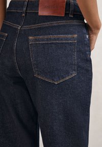 Detail tmavomodrých denimových džínsov zozadu so zašitými vreckami a hnedou koženou etiketou značky Reiss na páse.