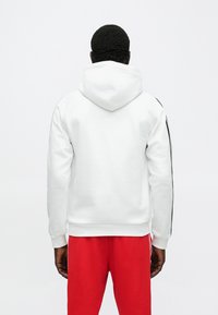Biela mikina s kapucňou s priliehavým strihom, vybavená veľkou kapucňou a čiernymi pruhmi na bokoch. Kombinovaná s červenými teplákmi, ktoré vytvárajú ležérny vzhľad.