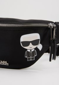 Zwarte nylon heuptas met zilveren rits, voorzien van een sticker van een cartoonfiguur in een zonnebril en pak aan de voorkant, gemarkeerd met "KARL."