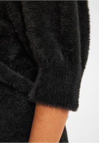 Sort fuzzy sweater med korte, brede ærmer og blød tekstur. Nærbillede fremhæver stoffets luksuriøse overflade og varme.
