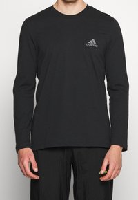 Mustat pitkät hiha t-paita, joka on valmistettu puuvillasta, pyöreällä pääntiellä ja hienovaraisella harmaalla Adidas-logolla vasemmassa rinnassa. Sujuva pinta.