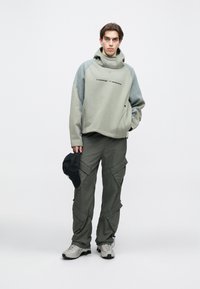 Ανοιχτό πράσινο πουλόβερ με ψηλό γιακά, γκρι λεπτομέρειες και λογότυπο, συνδυασμένο με σκούρα πράσινα cargo παντελόνια και γκρι αθλητικά παπούτσια. Κρατάει ένα μαύρο καπέλο.