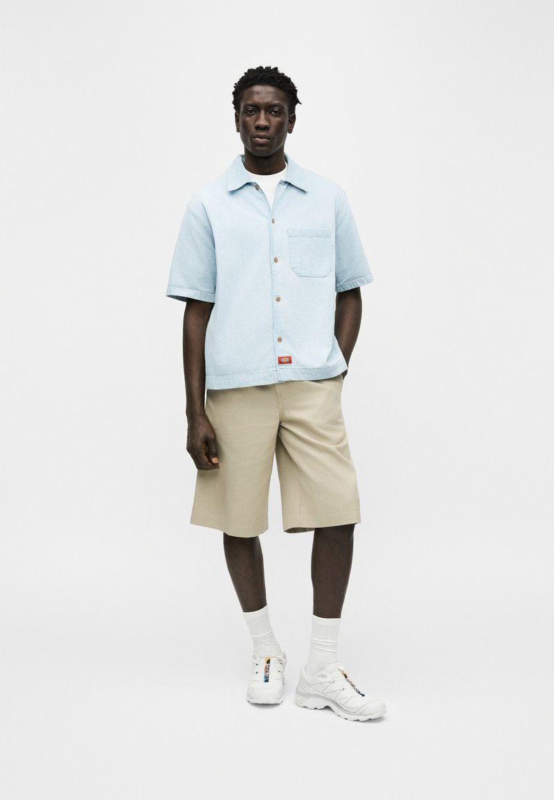 Homme debout portant une chemise à manches courtes bleu clair boutonnée, un short beige jusqu'aux genoux, des chaussettes blanches et des baskets blanches sur fond blanc.