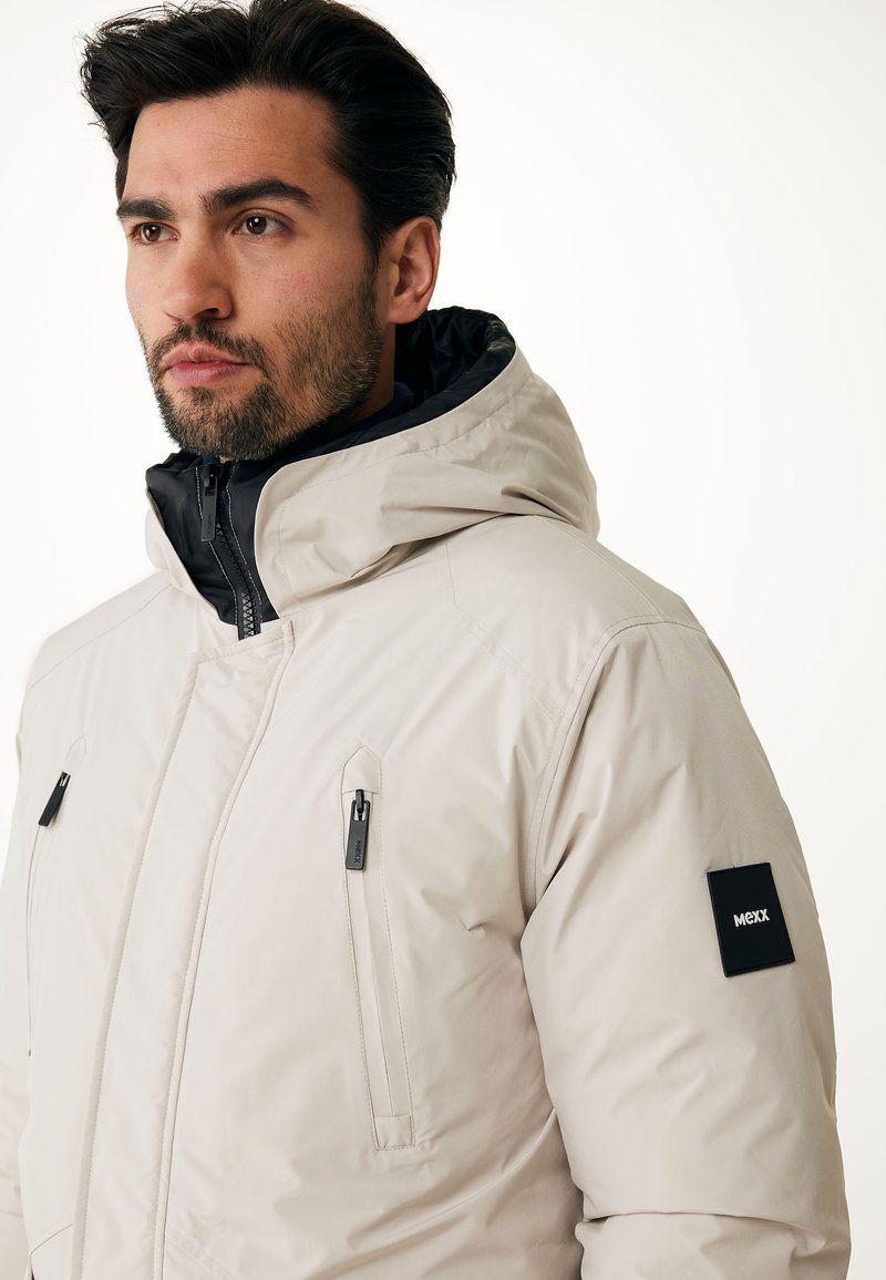 Chaqueta de un tono beige claro con un cuello alto y capucha. Presenta cremalleras y detalles en negro, dos bolsillos frontales con cremallera y un logo en la manga.