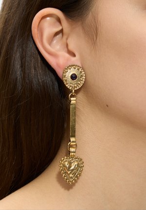 Złoty wiszący kolczyk z teksturowanym okrągłym sztyftem, ciemnym kamieniem w środku oraz zawieszką w kształcie serca wiszącą na prostokątnym łączniku na uchu kobiety.