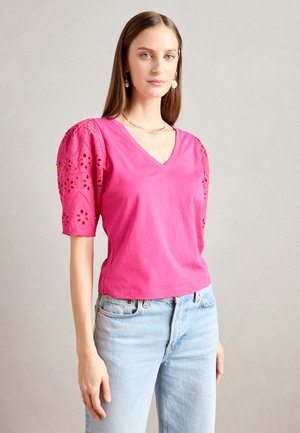 T-shirt en coton rose avec un col en V et des manches bouffantes, ornée de motifs floraux brodés sur les manches. Porté avec un jean bleu clair.