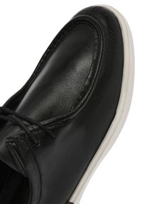 Chaussure en cuir noir avec une finition lisse, bout arrondi et semelle en caoutchouc blanche. Dispose de lacets et de détails de couture subtils le long du bord.