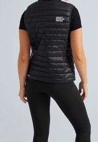 Czarna, pikowana kamizelka z zamkiem z przodu i poziomym przeszyciem. Posiada białe logo na plecach oraz dopasowany krój, noszona na czarne legginsy.