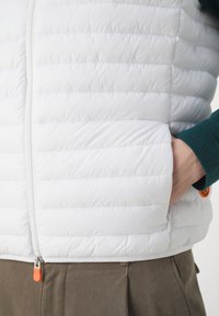 Hvid oppustelig vest med quiltet tekstur, lynlåslukning, sidelommer og orange detaljer. Bæres over en mørkegrøn top og beige bukser.