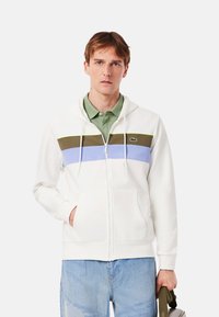 Sudadera blanca con cremallera y capucha, con franjas horizontales verdes, marrones y azules. Incluye un bolsillo frontal, capucha ajustable con cordón y detalle de logotipo.