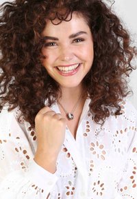 Kroes bruin haar, draagt een witte geborduurde blouse. Een zilveren ketting met een ronde hanger is zichtbaar, aangevuld met een glimlach.