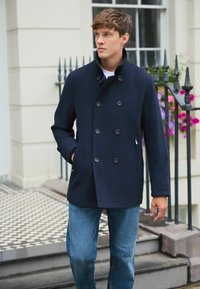 Marineblauer, doppelreihiger Mantel mit großen Knöpfen, Seitentaschen und hohem Kragen, kombiniert mit blauen Jeans. Strukturierter Stoff.