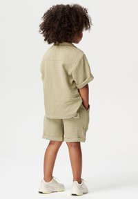 Khaki-textured short-sleeve shirt en bijpassende shorts, beide met opgerolde mouwen. Witte sneakers met gele accenten. Label zichtbaar op de shorts.