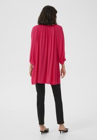 Blouse fuchsia à coupe amples, encolure froncée et manches trois-quarts, associée à un pantalon noir ajusté et des chaussures à petits talons.