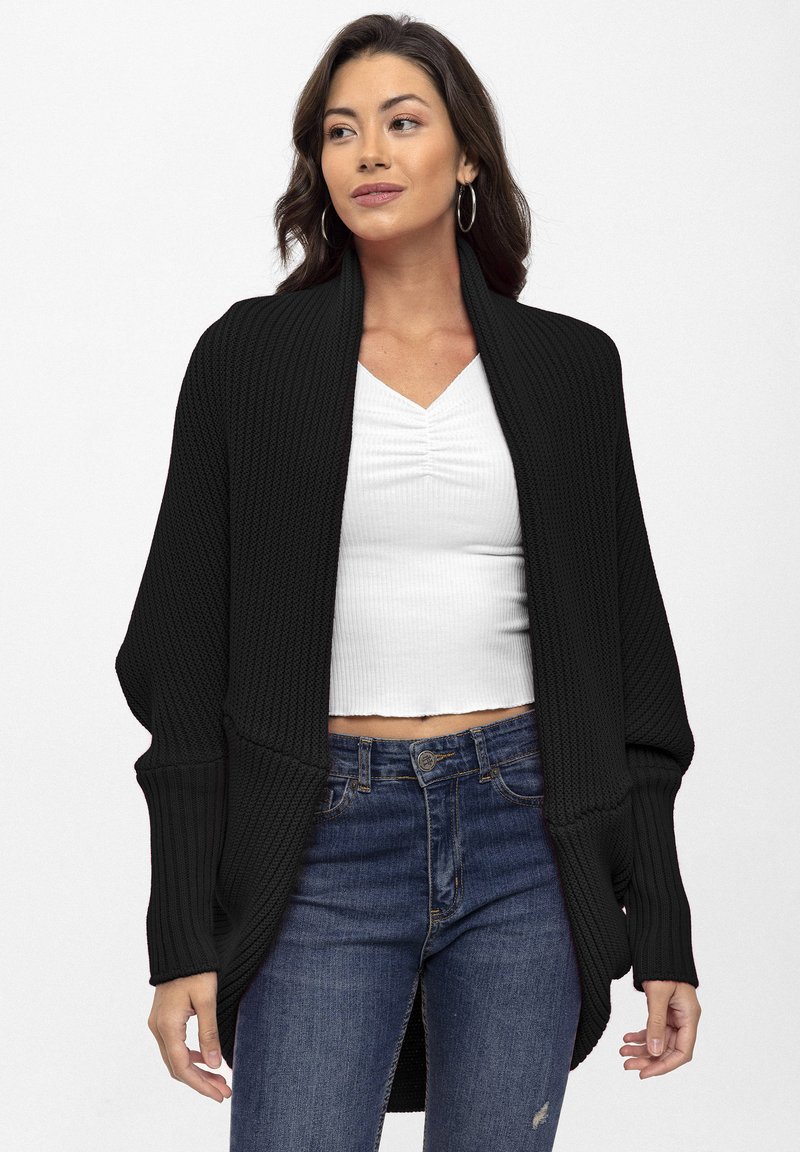 Cardigan nero a coste con apertura frontale e maniche lunghe, abbinato a un top bianco aderente e jeans blu, che mette in risalto il look casual a strati.