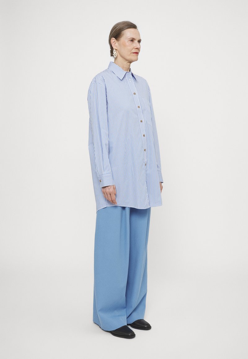 Blue and white striped button-up shirt with a collar, paired with wide blue trousers and black shoes. Simple, oversized fit.
