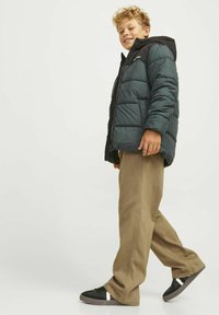Donkergroene pufferjack met een zwarte capuchon, voorzien van een ritsluiting, gewatteerde textuur en zijzakken, in combinatie met beige wijde broek en zwarte schoenen.