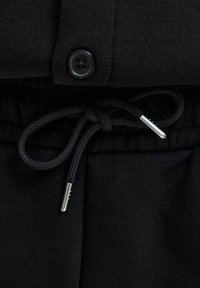 Tissu noir avec poignet boutonné au-dessus d'un cordon noir noué en nœud avec des embouts argentés à la taille d'un pantalon ou d'un short.