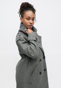 Harmaa villasekoitetakki, jossa on korkea kaulus, kaksirivinen malli, suuret mustat napit ja sivutaskut. Sileä rakenne, istuva leikkaus.