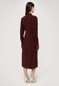Długa burgundowa sukienka o dopasowanej sylwetce, z zapięciem na guziki przy mankietach i gładką fakturą. Nosi ją w połączeniu z bordowymi rękawiczkami i kremowymi szpilkami.