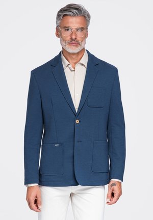 Hombre de mediana edad con cabello y barba canosos, que lleva gafas transparentes, blazer azul, camisa beige y pantalones blancos, de pie contra un fondo liso.