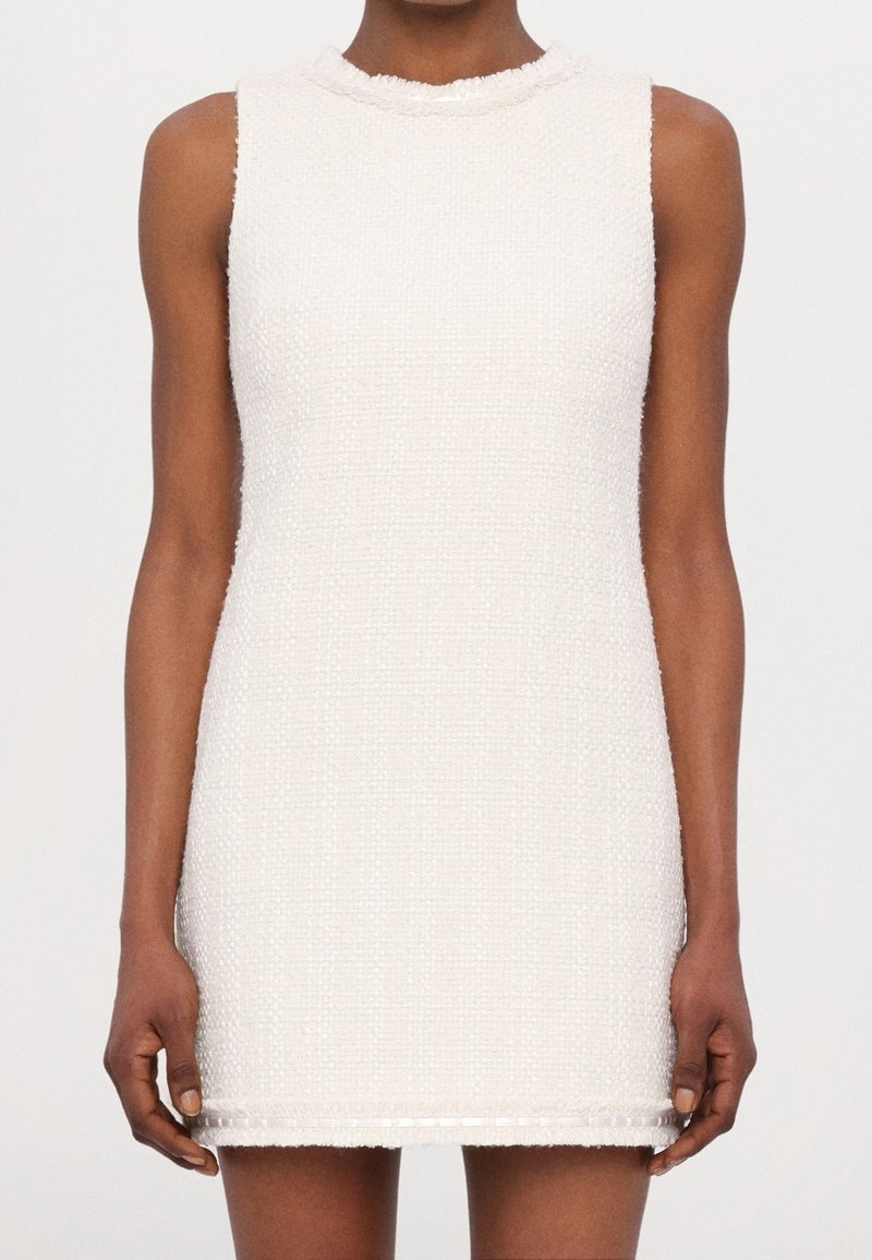 Robe mini blanche texturée sans manches avec un col rond haut portée par une personne sur un fond uni.