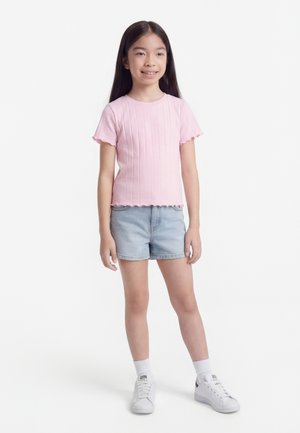 Κορίτσι με μακριά σκούρα μαλλιά, φορώντας ροζ κοντομάνικο μπλουζάκι, ανοιχτόχρωμα τζιν σορτς, λευκές κάλτσες και λευκά αθλητικά παπούτσια, στέκεται και χαμογελάει.