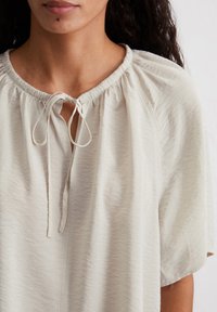 Helle beige Bluse mit gerafftem Halsausschnitt, kurzen Ärmeln und einer Schleifenverschlus. Hergestellt aus einem weichen, strukturierten Stoff mit lässiger Passform.
